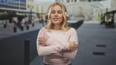 Kulaklık takan genç sarışın kadın şehir caddesinde durup sakin ve düşünceli bir şehir manzarası yaratırken kendini kucaklıyor..