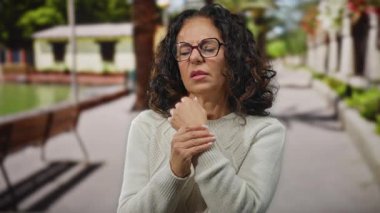 Kıvırcık saçlı, bileği ağrılı bir kadın güneşli bir sokakta..