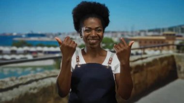 Güneşli bir sahil geçidine davetkar bir şekilde el kol hareketi yapan, önlük giyen, arka planda okyanus ve şehir manzarası olan bir kadın, davetkar bir açık hava ortamı yaratıyor..