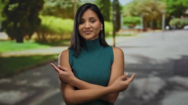 Deniz mavisi elbiseli genç İspanyol kadın güneşli park ortamında el kol hareketleri yapıyor. Arka planda ağaçlar ve patikalar olan rahat ve kendinden emin bir tavır sergiliyor..