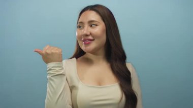 Açık mavi bir stüdyoda, bej bir bluz giyen ve uzun kahverengi saçlı bir kadın gülümsüyor ve başparmağıyla işaret ediyor..