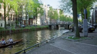 Parlak güneş ışığı altında yeşil ağaçlar ve tarihi binalar olan Amsterdam Kanalı 'nda kürek çeken adamların bulanık görüntüsü.
