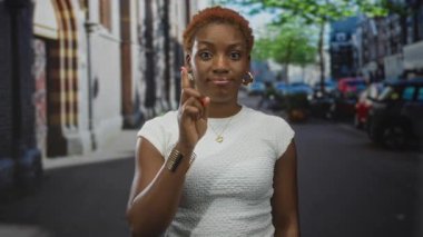Genç Afro-Amerikalı kadın, geçen arabalar ve mağazaların arasında sessiz şehir caddesinde sessiz kalmak için parmaklarını dudaklarına koyar; sessiz güven.