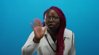 Canlı mavi arka plana karşı anlamlı jestlerle iletişim kuran, iletişimi ve duyguyu vurgulayan, gözlük takan ve çarpıcı bir görsel etki için örgülü saçlar takan bir kadın..