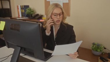Ofiste telefonla konuşan bir kadın, belgeleri incelerken, bilgisayar masası kurulmuş ve profesyonel kıyafetli verimli bir çalışma ortamı sergiliyor..