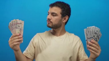 Suudi riyal ve Amerikan dolarlarını mavi stüdyoda karşılaştıran adam; finansal piyasa belirsizliği.