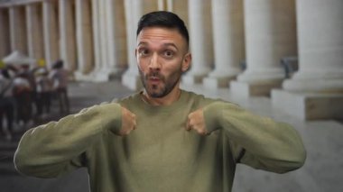 Eski üniversite kampüs koridorunda, günlük süveter jestleri yapan genç İspanyol adam merak ve nişanla....