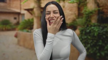 Açık havada gülümseyen genç bir kadın. Yüzünde beyaz bir tişört var. Etrafı koyu yeşil ve kahverengi..