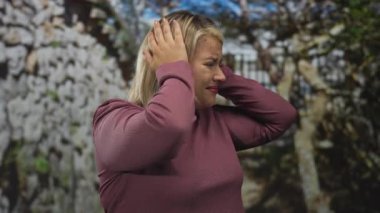 Sessiz bir şehir caddesinde taş bir kapının yanında elleri kulaklarının üzerinde tutan genç sarışın kadın rahatsız..