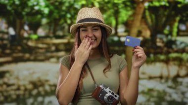 Parktaki İspanyol kadın, elinde kredi kartı ve neşeli bir ifadeyle ağaçlarla çevrili eski moda bir kamera tutuyor..