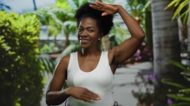 Güneşli bir parkta gülümseyen ve poz veren bir kadın. Etrafı yemyeşil, beyaz bir bluz giyiyor ve açık hava manzarasının tadını çıkarıyor..