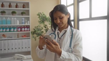 Doktor kıyafeti giymiş bir kadın klinik odasında telefona gülümsüyor. Parlak doğal ışık ve düzenli raflarla sıcak ve profesyonel bir sağlık hizmeti sunuyor..