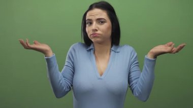 Mavi gömlekli kadın yeşil arka plana karşı belirsiz bir ifadeyle omuz silkiyor.