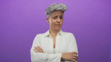 Kısa gri saçlı, büzüşmüş dudaklı, beyaz gömlek giyen ve mor bir stüdyoda kollarını kavuşturan beyaz bir kadın..