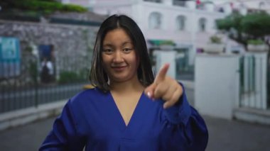 Mavi gömlekli, gülümseyen ve şehir arka planına sahip şehir caddesinde parlayan genç bir kadın, açık havada kendine güvenen ifadesini canlı bir şekilde vurguluyor..