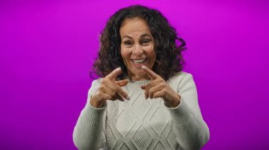 Kıvırcık saçlı olgun bir İspanyol kadın canlı mor bir arka planda jestler yapıyor, bir dizi anlamlı pozlarda sıcaklığını ve yakınlığını sergiliyor..