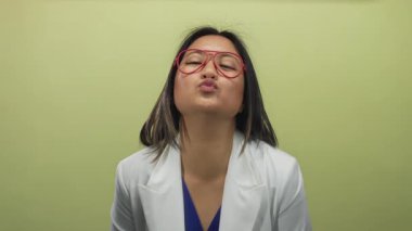 Beyaz ceketli ve kırmızı gözlüklü genç bir kadın kameraya bir öpücük veriyor ve izole edilmiş sarı arka plana karşı elleriyle kalp hareketi yapıyor..