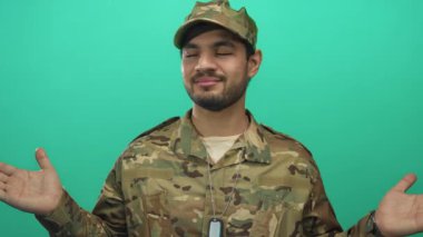 Asker üniformalı adam stüdyoda yeşil arka plana karşı açık el hareketi gösteriyor; belirsizlik.