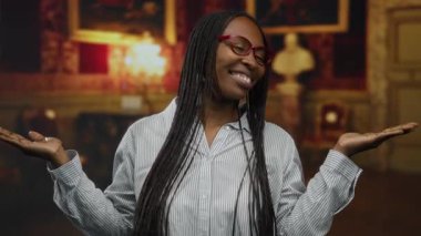 Kapalı müze ortamında kollarını açmış gülümseyen bir kadın, gözlüğünü ve örgülü saçlarını zarif bir arka planda örüyor..