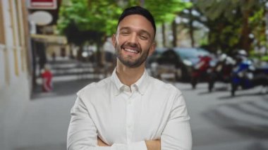 Beyaz gömlekli genç İspanyol adam kollarını kavuşturuyor ve arka planda bulanık ağaçlar ve scooterlarla hareketli bir şehir caddesinde gülümsüyor, canlı şehir yaşamını kucaklıyor..