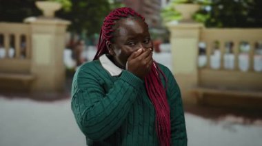 Şehir caddesinde ağzı kapalı bir şekilde gülen bir kadın yeşil bir kazak ve pembe örgülü saçlarla şehir ortamını ve güneş ışığını gösteren bulanık bir arka planı var..