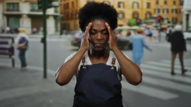 Önlük giymiş bir kadın şehir caddesinde sakin bir şekilde duruyor. Arka planı bulanık, şehir dışı hareketlerini tasvir ediyor..