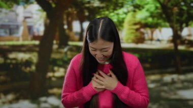 Canlı pembe bir kazakla gülümseyen kadın güzel yeşil bir parkta güneşli bir günün tadını çıkarıyor. Mutluluğunu doğa ve ağaçlarla ifade ediyor. Açık havada neşeli anlar sergiliyor..