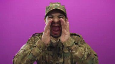 Kamuflaj üniformalı genç İspanyol bir adam mor bir stüdyoda elleri çerçeveli bağırıyor..