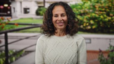 Şehir caddesinde gülümseyen bir kadın, mutluluk ve samimiyet gösteriyor, kıvırcık saçlı, rahat bir kazak giyiyor ve yeşilliklerle çevrili..