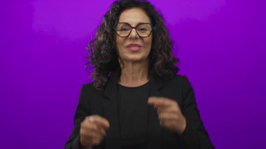 Siyah ceketli orta yaşlı İspanyol kadın mor stüdyo duvarına gülümsemek için işaret ediyor..