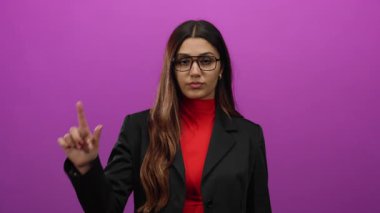 Gözlüklü genç İspanyol kadın pembe arka planda ezik jestler yapıyor, kırmızı ve siyah elbiseler giyiyor, ciddi bir yüz ifadesi sergiliyor..