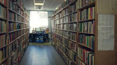 Sessiz bir kütüphanenin bulanık görüntüsü, kitapların sergilendiği satırlar ve çalışma masasının yumuşak odak noktası.