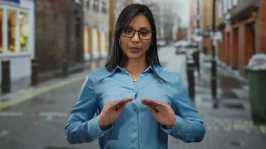 Mavi gözlüklü kadın şehir sokaklarında el kol hareketi yapıyor. Genç ve odaklanmış. Açık şehir ortamında sakin ve anlayışlı..