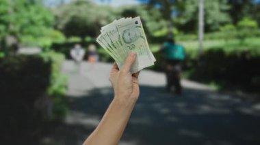 Güneşli bir caddede elinde Singapur doları tutan adam şehirde para ve açık hava aktivitelerini ön plana çıkarıyor..
