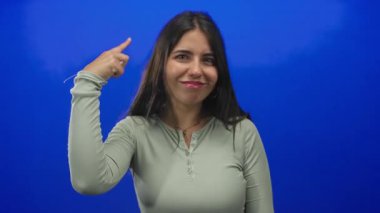 Uzun saçlı genç kadın, yeşil uzun kollu bluzuyla gülümserken stüdyoda mavi duvarlı parmak ucuyla tapınağa işaret ediyor..