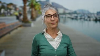 Üst düzey bir kadın kıyı limanında gri saçlarıyla, arka planda tekneleriyle güzel bir jest yapıyor. Açık hava anının tadını çıkarıyor. Gözlüklerine odaklanıyor ve gülümsüyor..