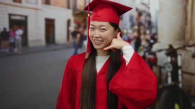 Kırmızı mezuniyet elbisesi giymiş bir kadın, şehir sokağında akademik başarıya güveni gösteren neşeli bir ifadeyle çağrı yapıyor.