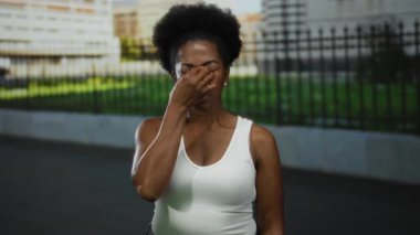 Beyaz kolsuz tişörtlü kadın, arka planda binalar olan şehir caddesinde yorgun görünen burnunu tutuyor..