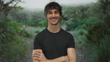 Siyah t-shirt giyen ve kolları çapraz duran bir adam yeşil orman yolu üzerinde açık hava ayarı; memnuniyet.