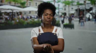 Açık şehir restoranının terasında duran önlük giymiş bir kadın bulanık sokak geçmişiyle şehir çalışma ortamını yakalıyor..