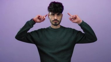 Koyu renk saçlı genç bir adam kulaklarını kapatırken, iç mekanda mor bir arka plana karşı rahatsızlığını dile getiriyor.