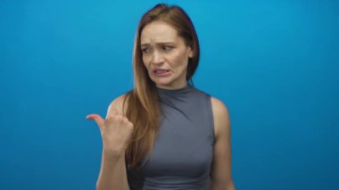 Mavi stüdyo ortamında kolsuz, gri tişörtlü, dramatik başparmağıyla kaşlarını çatan bir kadın..