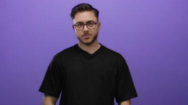 Sakallı, gözlüklü, parmakları mor arka planda selam veren, günlük siyah gömlek giyen, stüdyo ortamında dostane ve cana yakın bir jest yapan genç bir adam..