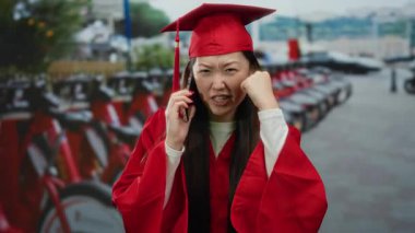 Kırmızı mezuniyet elbisesi giymiş bir kadın, arka planda motosikletlerle şehir sokağında telefonla konuşurken öfkeyle el sallıyor..