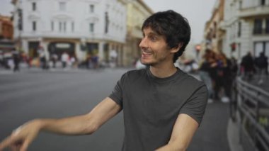 Yoğun şehir sokaklarında genç bir adam her iki işaret parmağını çevreleyen binalara ve mağaza vitrinlerine neşeli bir sırıtışla işaret ediyor..