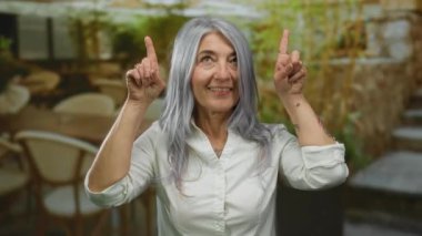 Açık hava restoranının terasında beyaz saçlı yaşlı bir kadın. Sokak manzaralı, canlı bir enerjisi var..