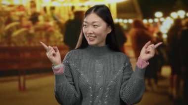 Kadın gülümsüyor ve açık hava fuarına işaret ediyor. Arka planda parlak ışıklar var. Canlı bir akşam atmosferinde..