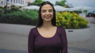 Açık havada duran bir kadın kentin yeşilliği içinde kendine güvenen bir gülümsemeyle ve parlak mor gömleğiyle arka planı bulanık bir şekilde pozitif enerji ve gençlik güvenini yakalıyor..