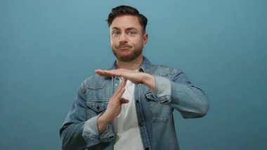 Sakallı genç adam izole edilmiş mavi arka plana karşı jest yapıyor, kot gömlek giyiyor, mola ya da duraklama ihtiyacını ifade ediyor, duygularını yüz ifadeleriyle ifade ediyor ve el işaretleriyle ifade ediyor..
