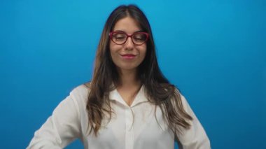 Gözlüklü ve beyaz gömlekli bir kadın canlı mavi arka plana karşı güvenle gülümsüyor, izole edilmiş bir ortamda modern İspanyol güzelliğini sergiliyor..